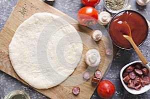Top view of raw dough for pizza on the cutting board and various ingredients for it.Tomatoes, sauce, mushrooms, oil, herbs, sausag