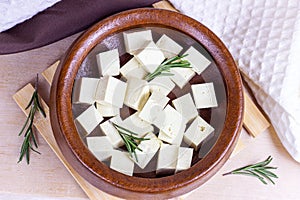 Top view of raw organic vegetarian tofu cubes in round bowl with fresh rosemary on wooden background.