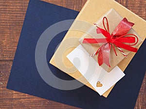 Top view, red bow tie brown gift box and heart-shaped punching white card over blue paper on wooden background.