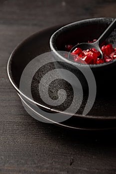 Top view of red pomegranate seeds in bowl with dark spoon and black dishes, on dark wooden table