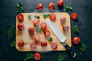 Top view of red sliced tomatoes on wooden chopping board. Sharp knife near. Green parsley and dill. Dark background. Preparing