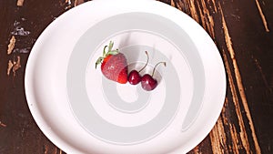 Top view of red strawberry and two sweet red cherries in the white plate on a wooden background. Space for text. Design concept