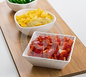 Top view of red, yellow, and peppers cut into cubes in white saucers on a wood cutting