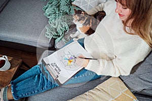 Top view relaxed young woman drawing work-life balance wheel sitting on the sofa with cat pet at home. Self-reflection