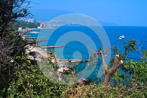 top view of resort town with white buildings on shore of Black Sea bay among mountains, blue water, green forest, broken trees