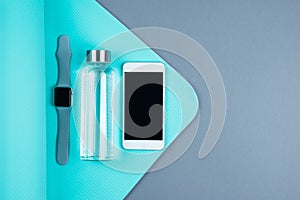 Top view of reusable eco glass water bottle, smart mobile phone and tracker watch on turquoise blue textured yoga mat