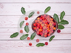 Top view of ripe red acerola cherries fruit in a ceramic bowl and green leaves on a wooden table.