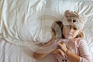 Top view of cute blonde little girl in pajamas and blindfold lying in white bed, awaking early in the morning before going to