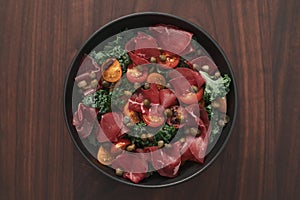 top view of salad with kale, bresaola and cherry tomatoes decorated with capers in black bowl on walnut wood table