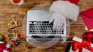 Top view Santa hands typing on keyboard and drinks tea by wooden decorated table. Santa Claus works with laptop, looks