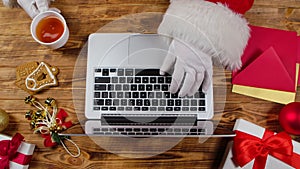 Top view Santa hands are typing on keyboard, drinks tea by wooden decorated table. Santa Claus works with laptop, looks