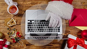 Top view Santa hands are typing on keyboard, drinks tea by wooden decorated table. Santa Claus works with laptop, looks