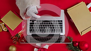 Top view Santa hands in white gloves are typing on keyboard laptop by red New Year decorated table. Santa Claus looks