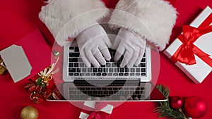 Top view Santa hands in white gloves are typing on keyboard laptop by red New Year decorated table. Santa Claus looks