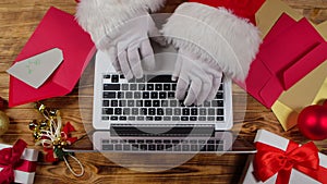 Top view Santa hands in white gloves are typing on keyboard laptop by wooden New Year decorated table. Santa Claus looks