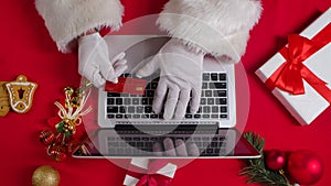 Top view Santa hands in white gloves are typing on the keyboard by red New Year decorated table. Santa Claus works with