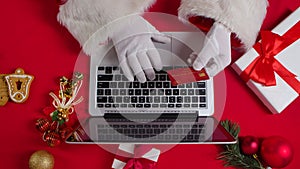 Top view Santa hands in white gloves are typing on the keyboard by red New Year decorated table. Santa Claus works with