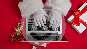 Top view Santa hands in white gloves typing on the keyboard by red New Year decorated table. Santa Claus works with