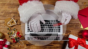 Top view Santa hands in white gloves are typing on the keyboard by wooden New Year decorated table. Santa Claus works