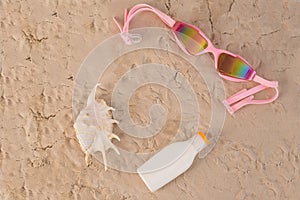 Top view of seashell swimming goggles and sunscreen lotion on sand.