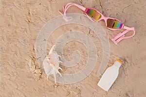 Top view of seashell swimming goggles and sunscreen lotion on sand.