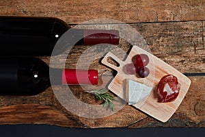 Top view of served snacks on board with twig of rosemary and bottles of red wine on wood