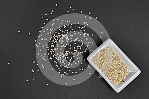 Top view of sesame seeds in a little dish and spilled on gray paper