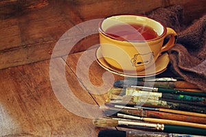 Top view of set of used paint brushes, cup of hot tea and palette over wooden table