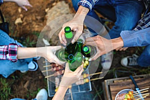 Top view shot of friends clinking bottle of beer during camping outdoor with barbecue on grill below in background. Celebrating