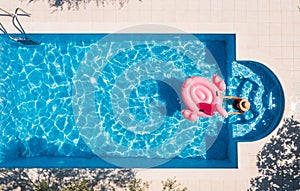 Top view shot of a woman in a straw hat with an inflatable pink flamingo entering a blue water swimming pool. Summer vacation,