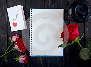 top view of a sketchbook and red color roses with a postcard and paper cup of coffee on dark wooden background