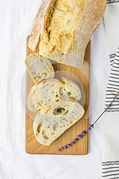 Top View of Sliced Artisan Rustic Bread on Wooden Cutting Board White Line Table Cloth Lavender Twig. Golden Crust