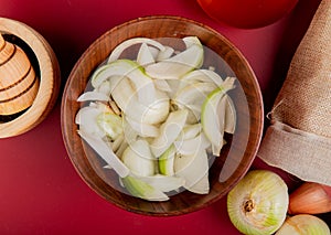 top view of sliced white onion in bowl with whole ones spilling out of sack and black pepper in garlic crusher on red background