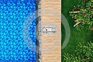 Top view of slim young woman in white bikini and straw hat lying on towel near swimming pool. Back view, without face