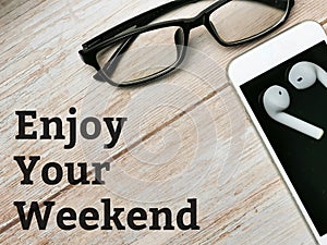 Top view smartphone,eye glasses and ear buds with text ENJOY YOUR WEEKEND written on wooden background