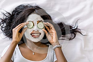 Top-view of smiling African woman making spa procedures with mud facial mask and fresh cucumber slices on eyes, laying