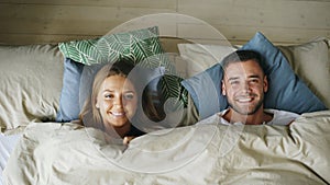 Top view of smiling couple having fun in bed hiding under blanket and looking into camera