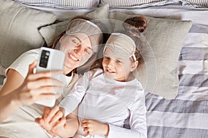 Top view of smiling happy family waking up in morning lying in bed in sleeping masks and making selfie woman holding smart phone