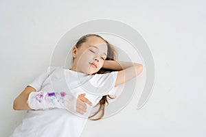 High-angle view of positive sweet little girl with broken hand wrapped in plaster bandage with colorful draw lying on