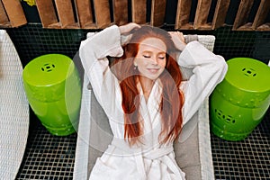 Top view of smiling young woman in white bathrobe relaxing with closed eyes lying on deck chair after beauty treatment