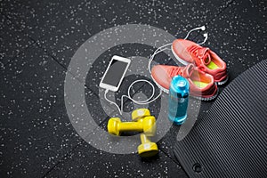 A top view of sportive accessories for gym training. Sport shoes, dumbbells, bottle, and a white phone on a black