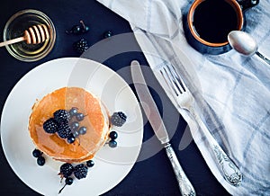 Top view stack of pancakes with blackberry and black currant, cup of coffee and saucer with honey on dark background.