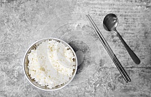 Top view steamed rice in a stainless bowl with spoon and chopsticks on the gray concrete table