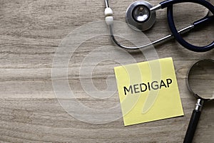 Top view of stethoscope, magnifying glass and yellow paper note written with Medigap on wooden background with copy space