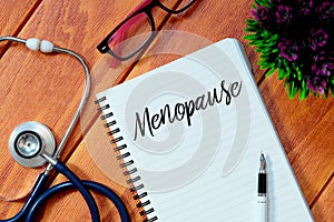 Top view of stethoscope,pink eyeglasses,plant,pen and notebook written with Menopause on wooden background.Health concept.