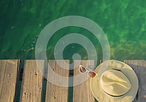 Top view of straw hat and sunglasses on wooden bridge over emerald green sea water. Summer vacation travel background. Summer vibe