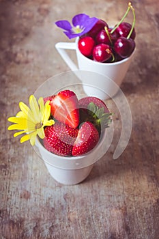 Top view of strawberries, cherries and yellow and purple flowers in bowls on wooden background