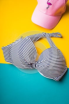 Top view of striped blue and white swimming bra and pink beach hat with flamingo on blue and yellow pastel background