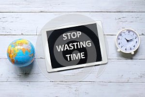 Top view of table clock,globe and tablet written with message Stop Wasting Time on wooden background.
