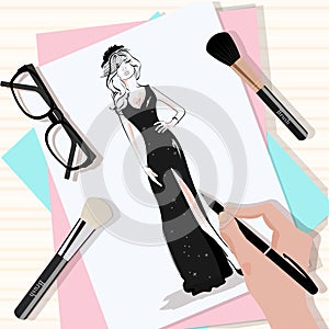 Top view of the table with papers, hand drawn fashion woman in black dress, brushes, eyeglasses and hand with pen.